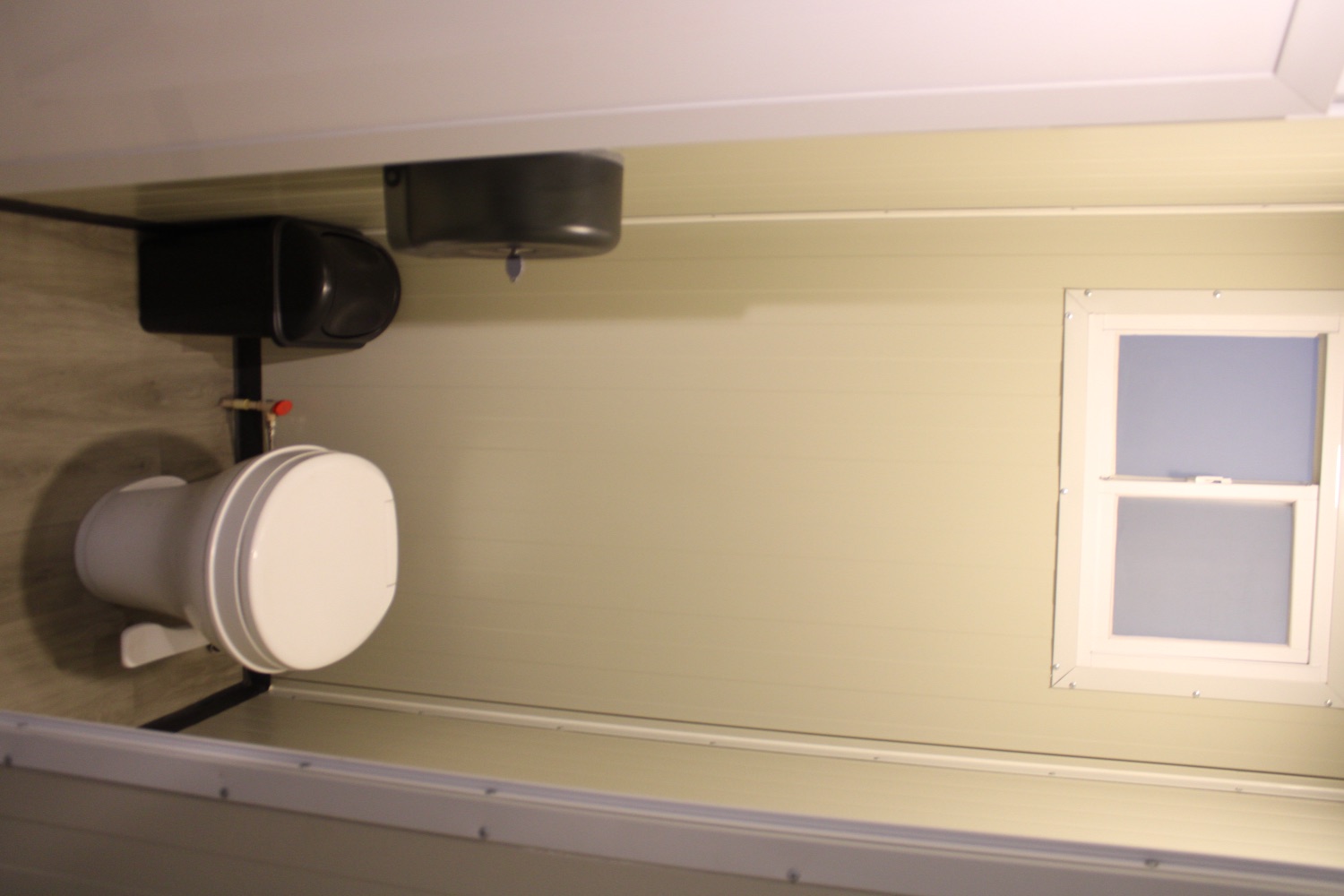Sanitario individual con ventana, dispensador de papel y piso de madera en el interior de la unidad